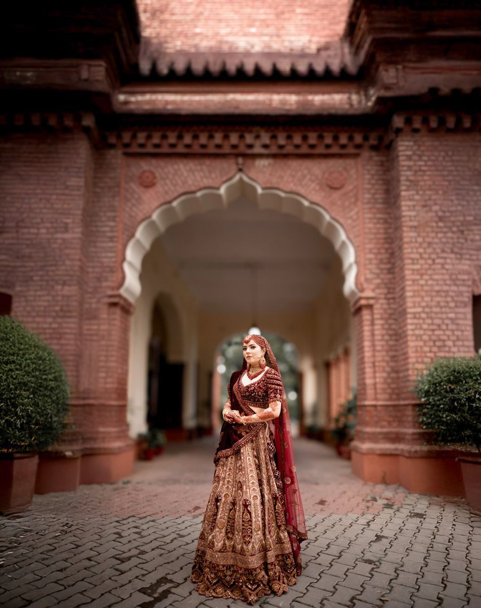 Bride in a maroon and gold embellished lehenga standing beneath a red sandstone Mughal archway, Adam & Sobia's wedding at Chamba House, Lahore