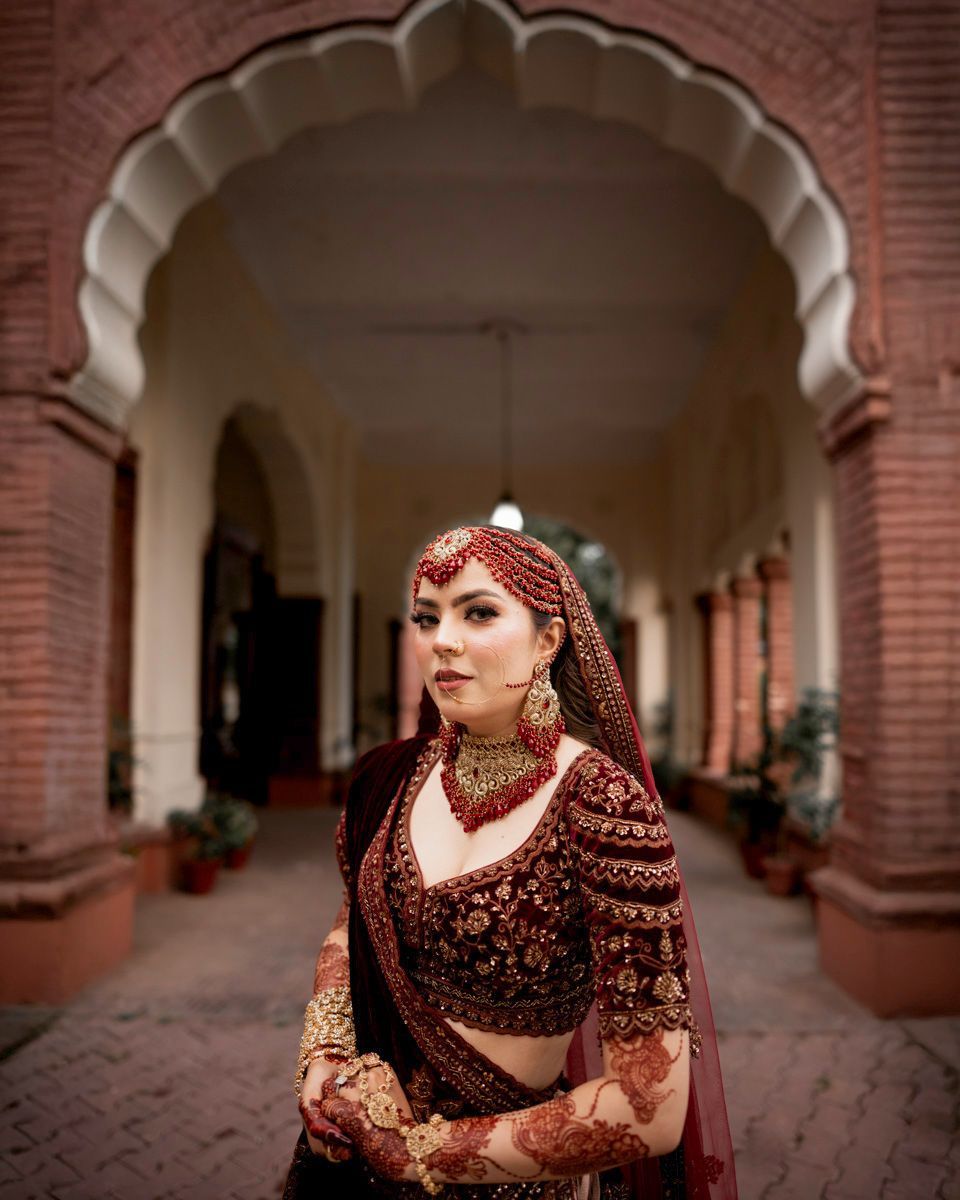 Close-up bridal portrait beneath the brick colonnade, wearing a red matha patti and gold jewelry, Adam & Sobia's wedding at Chamba House, Lahore