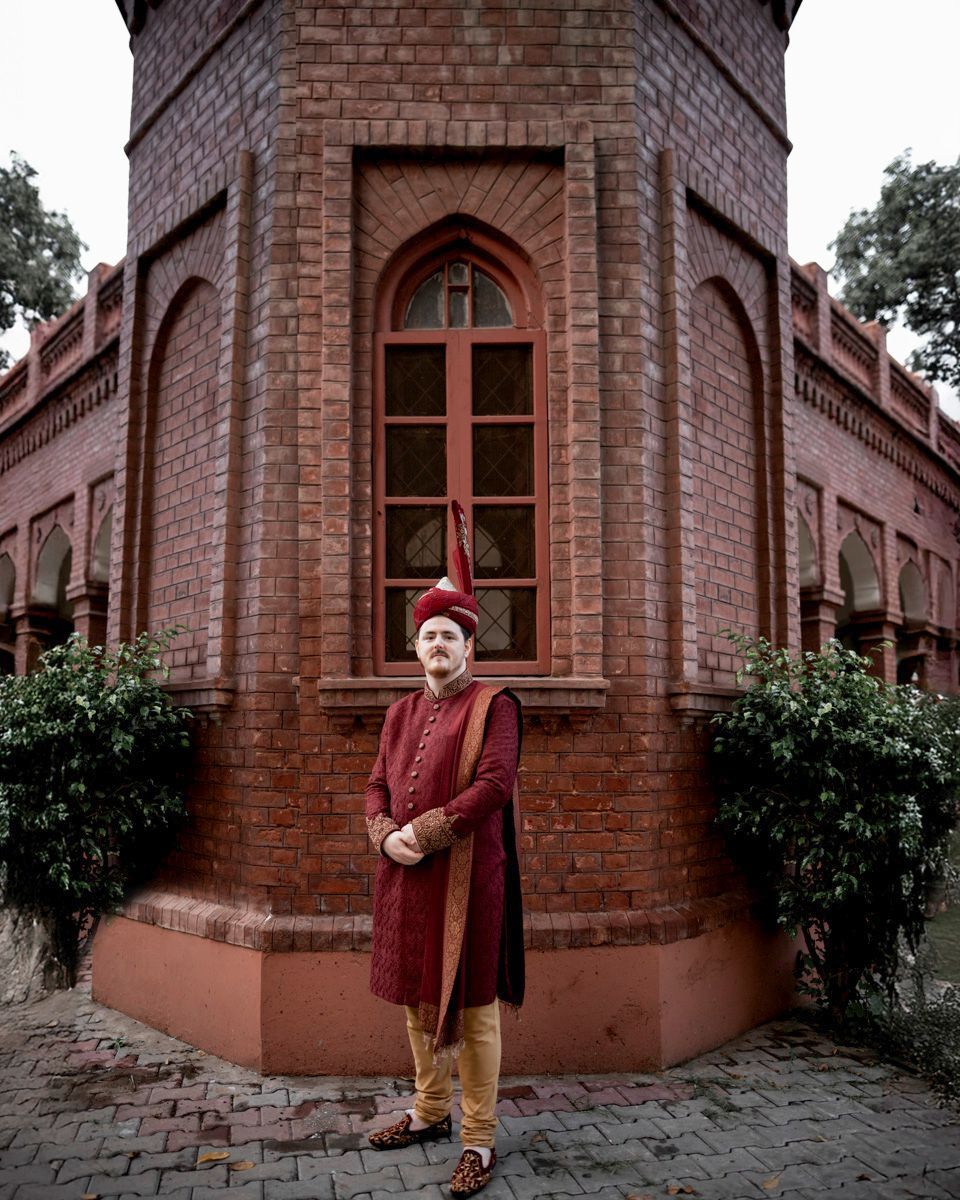 Solo groom portrait in a maroon sherwani and turban with a tall kalgi, standing before an octagonal brick tower, Adam & Sobia's wedding at Chamba House, Lahore