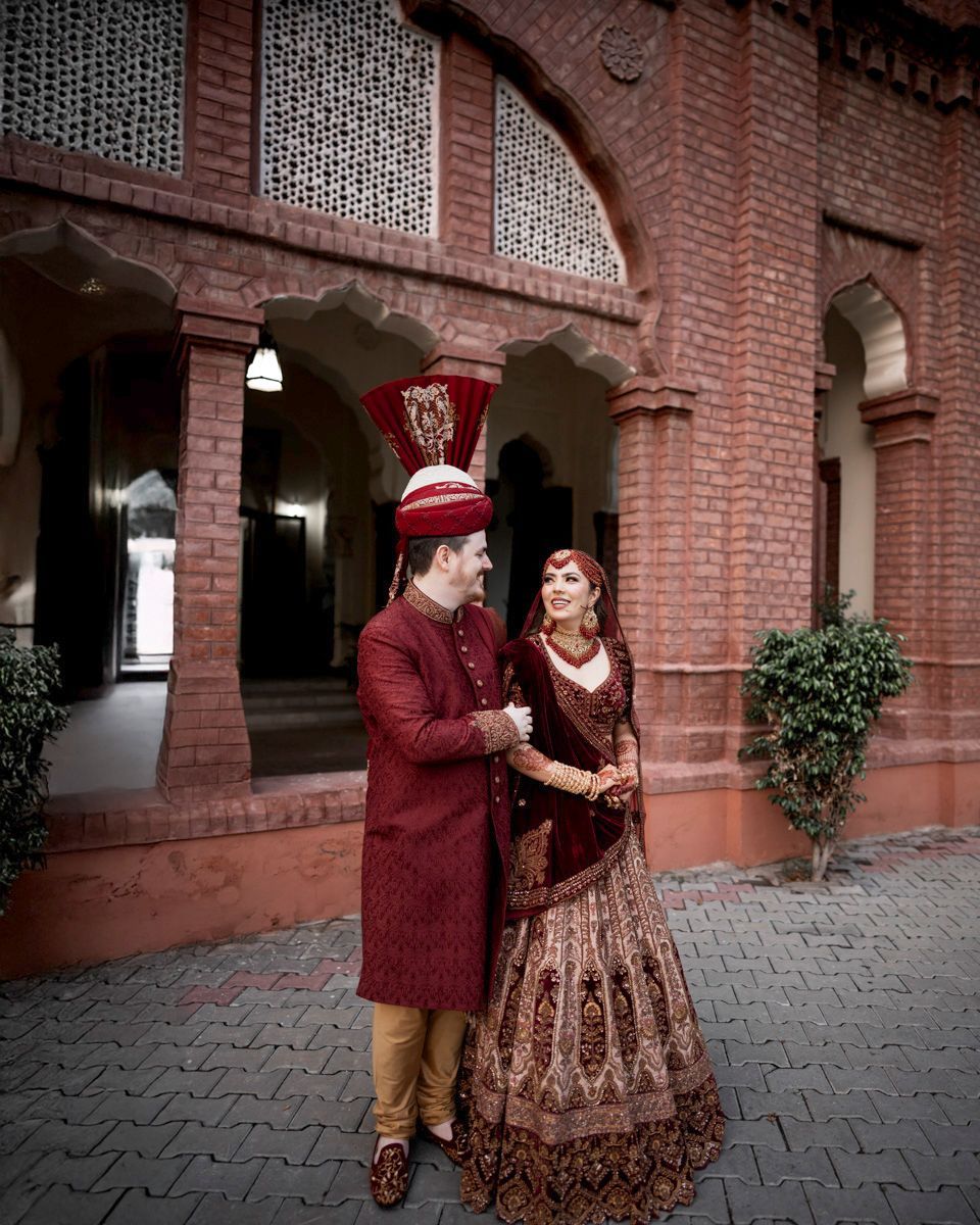 Bride and groom standing together for the first time in matching maroon outfits in front of the honeycomb-patterned brick facade, Adam & Sobia's wedding at Chamba House, Lahore