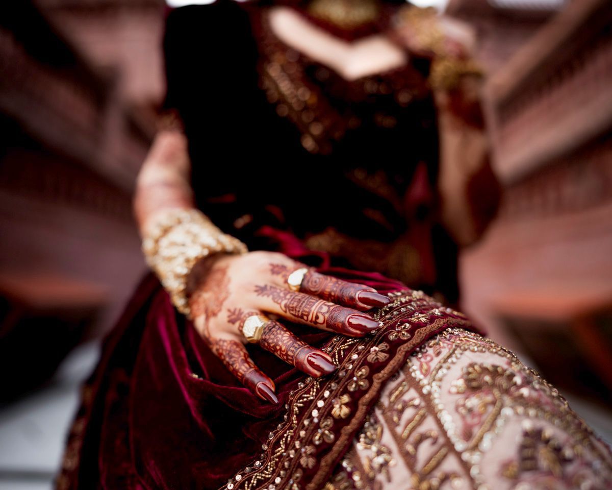 Detail of the bride's henna-painted hand resting on her lehenga skirt, brick towers blurred behind, Adam & Sobia's wedding at Chamba House, Lahore