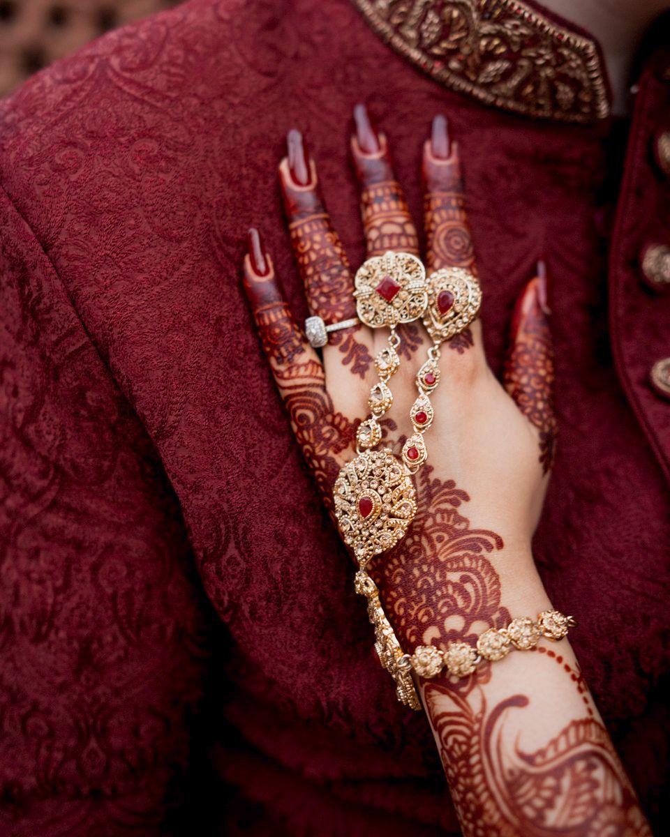 Detail of the bride's henna-painted hand and gold rings against her maroon sherwani-clad partner, Adam & Sobia's wedding at Chamba House, Lahore