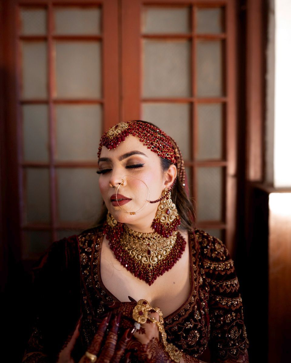 Solo bridal portrait indoors with eyes closed, wearing a red beaded matha patti, wooden doors behind her, Adam & Sobia's wedding