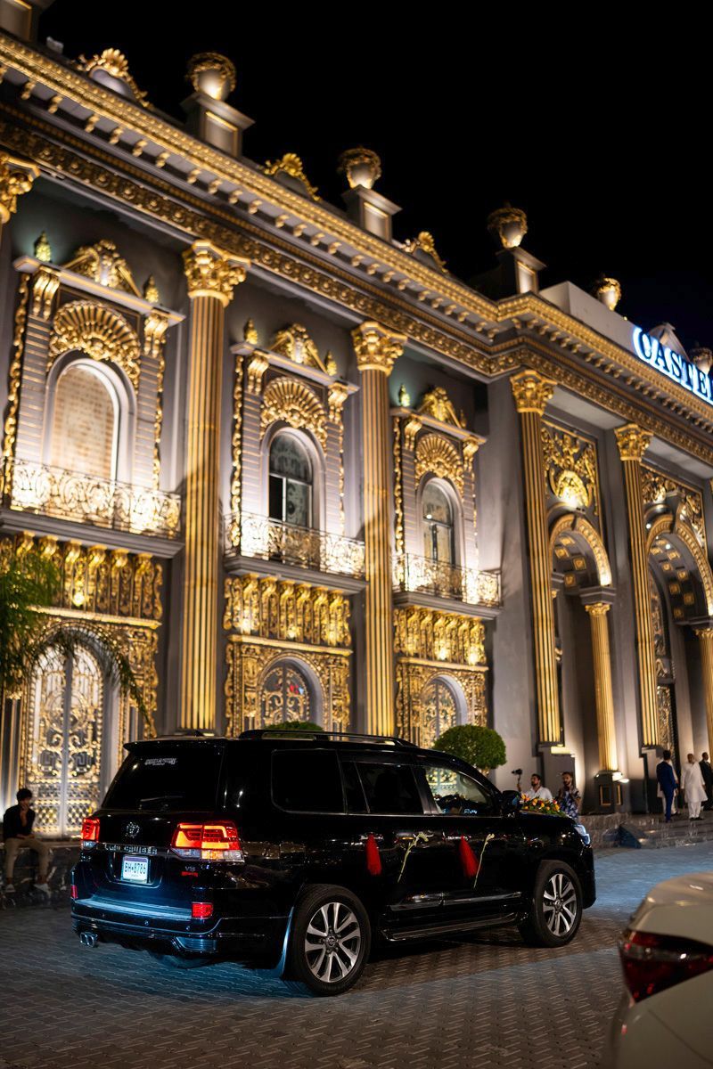 A flower-decorated SUV parked outside the illuminated gold-columned reception venue at night, Adam & Sobia's wedding