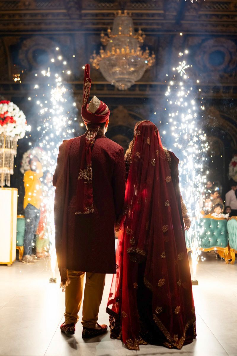 Bride and groom seen from behind walking through indoor cold-spark fireworks beneath a crystal chandelier at their grand entrance, Adam & Sobia's wedding