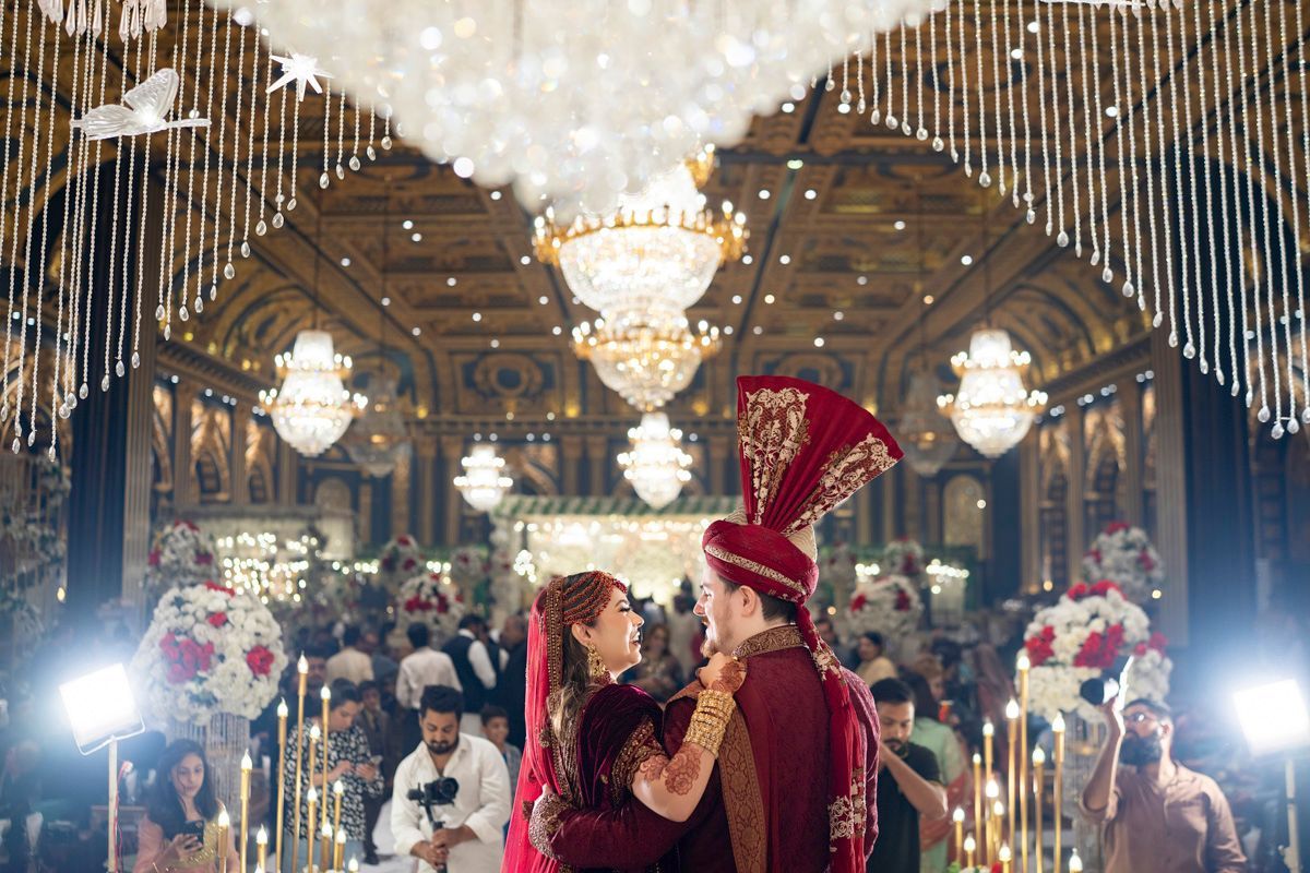 Bride and groom embracing on the dance floor beneath ornate chandeliers, guests and photographers visible around them, Adam & Sobia's wedding