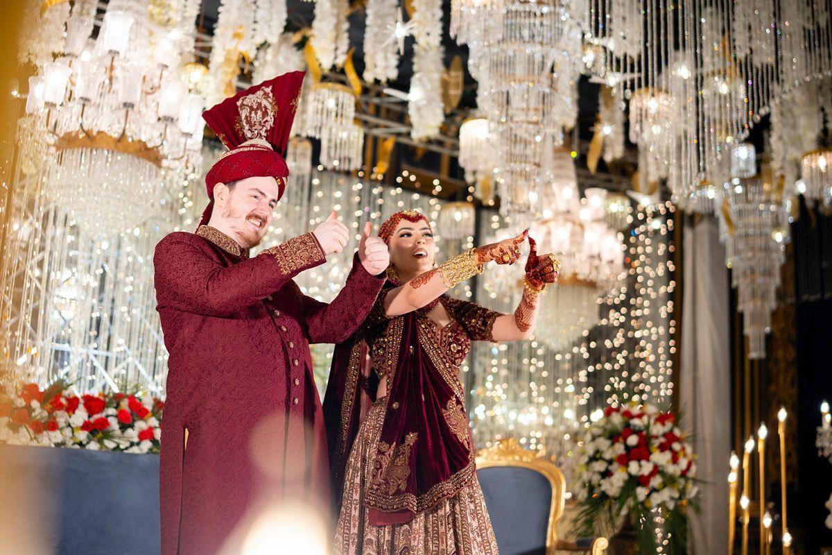 Bride and groom giving a thumbs-up and pointing at the camera, surrounded by hanging crystal chandeliers, Adam & Sobia's wedding