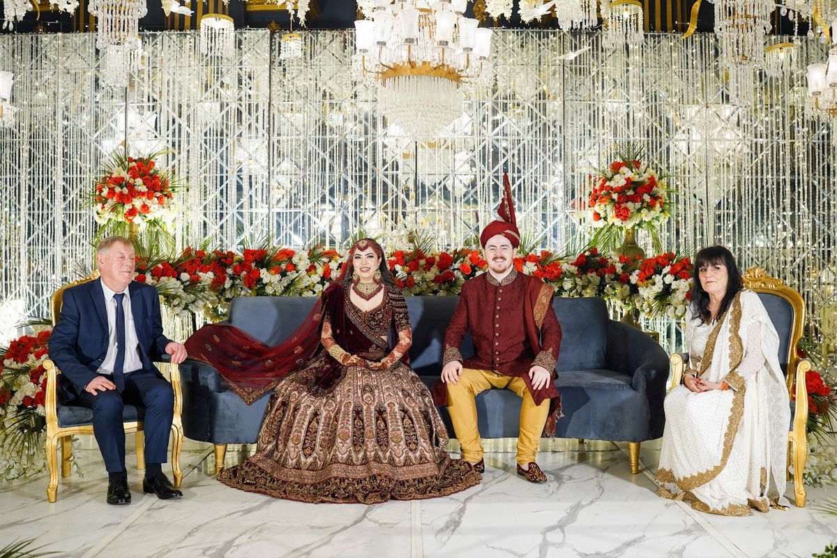 Bride and groom seated on a stage sofa with the groom's parents beside them, floral and crystal backdrop, Adam & Sobia's wedding