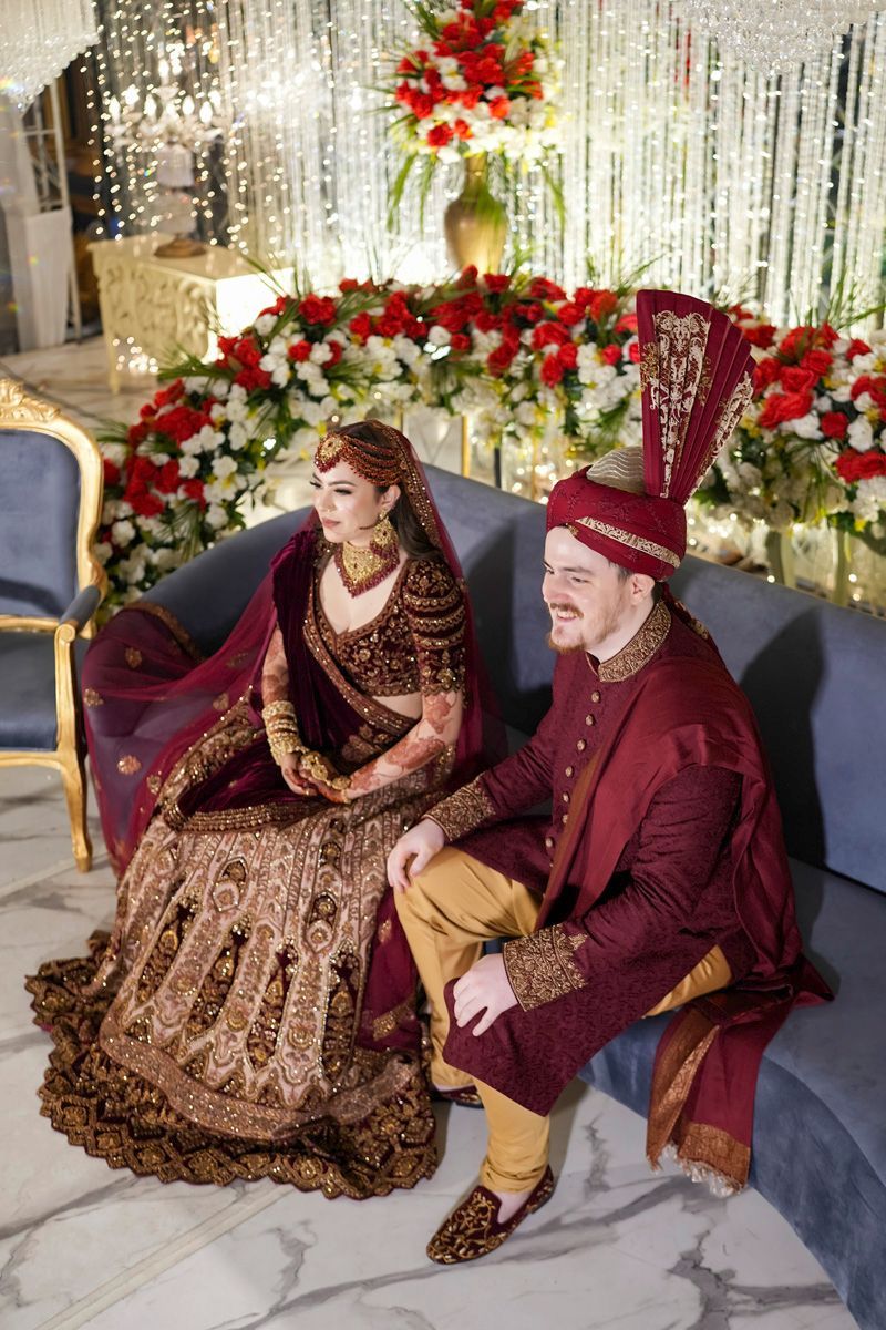 Bride seated and groom kneeling beside her on a stage sofa, floral and crystal curtain backdrop, Adam & Sobia's wedding