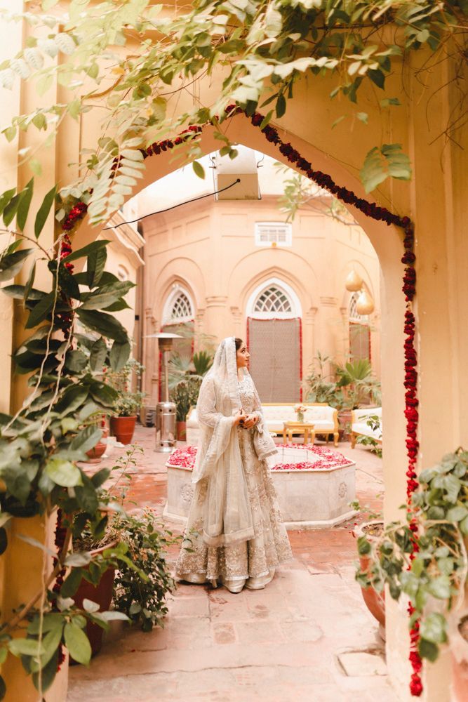 Bride viewed through a flower-draped archway, standing beside the haveli's courtyard fountain, Ahmed & Laraib's wedding at Barood Khana Haveli, Lahore