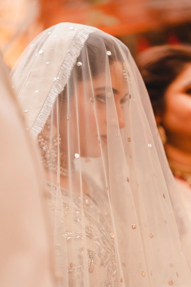 Close-up profile portrait of the bride beneath her sequined veil, a guest blurred behind her, Ahmed & Laraib's wedding