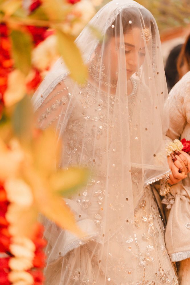 Bride beneath her veil holding a floral wristlet, framed by hanging red and cream garlands, Ahmed & Laraib's wedding