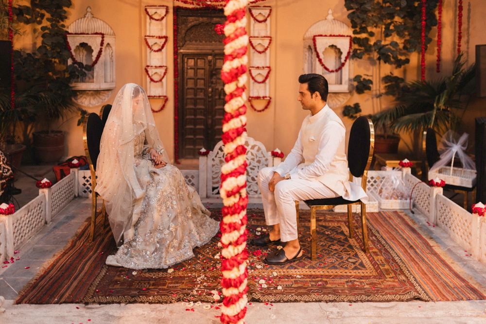 Bride and groom seated for their nikah ceremony in the haveli courtyard, separated by a red and white floral pillar, Ahmed & Laraib's wedding at Barood Khana Haveli, Lahore