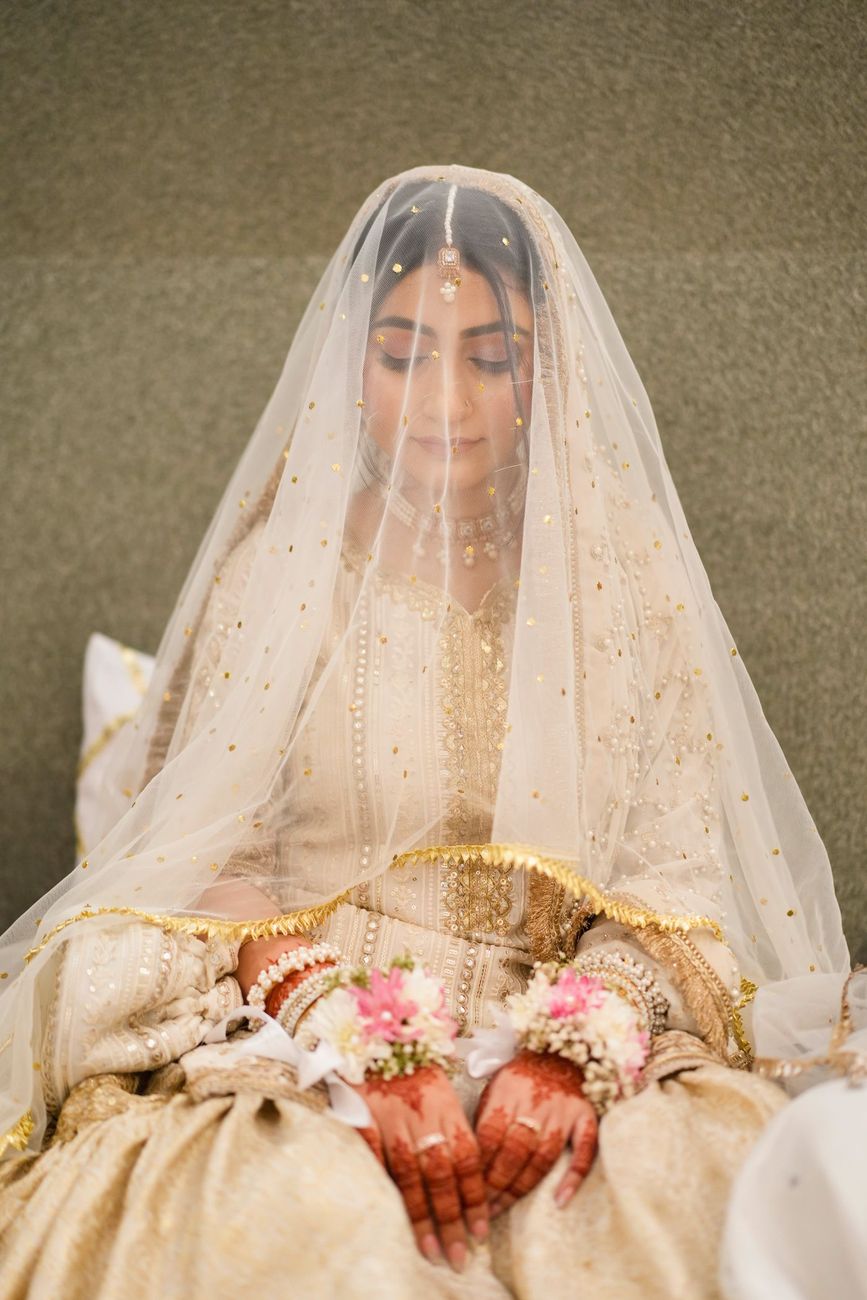 Bride seated beneath her veil holding a floral wristlet, eyes closed in quiet reflection before the nikah, Maham & Aurangzeb's wedding