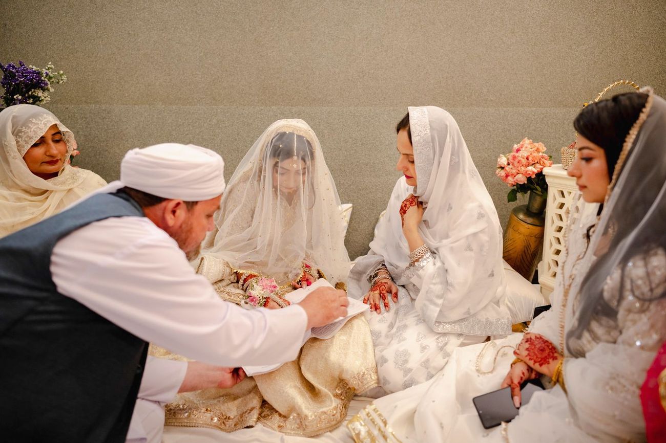 Bride signing the nikah register as the imam writes and family look on, Maham & Aurangzeb's wedding
