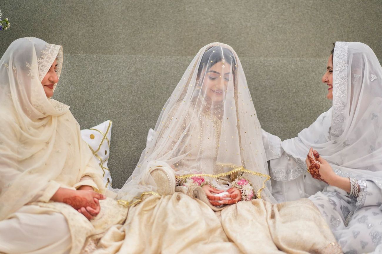 Bride seated beneath her veil flanked by two female relatives sharing a smile, Maham & Aurangzeb's wedding