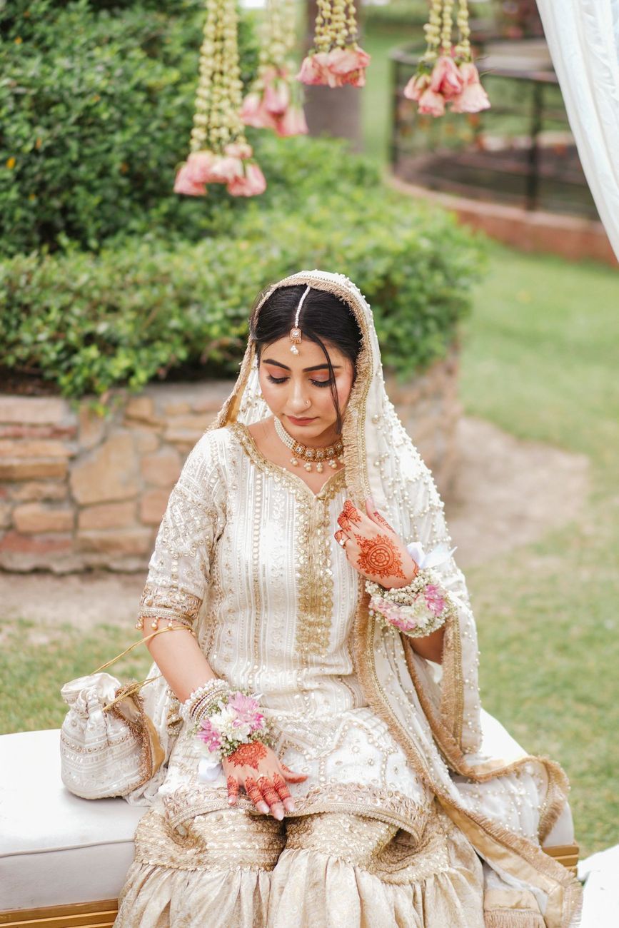 Solo bridal portrait seated outdoors in a garden with hanging floral garlands above her, Maham & Aurangzeb's wedding