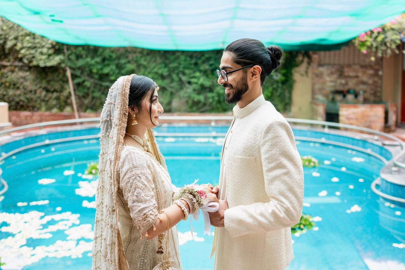 Bride and groom holding hands beside a pool scattered with flower petals, under a teal shade canopy, Maham & Aurangzeb's wedding