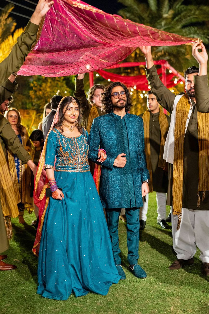 Bride and groom in matching teal mehndi outfits walking beneath a pink dupatta canopy held by friends, Maham & Aurangzeb's wedding
