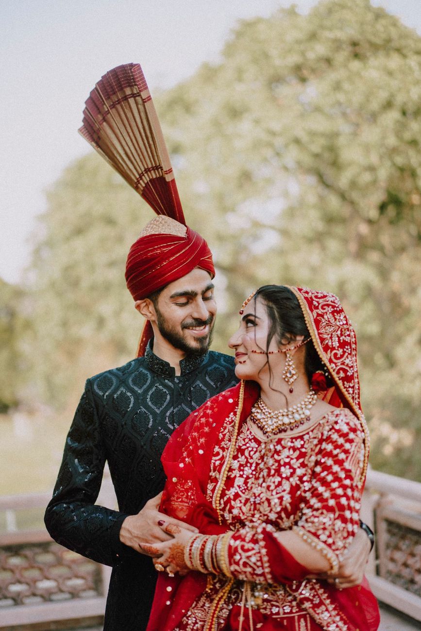 Close-up of bride and groom smiling at each other, groom in a black sherwani and red turban, Maham & Aurangzeb's wedding