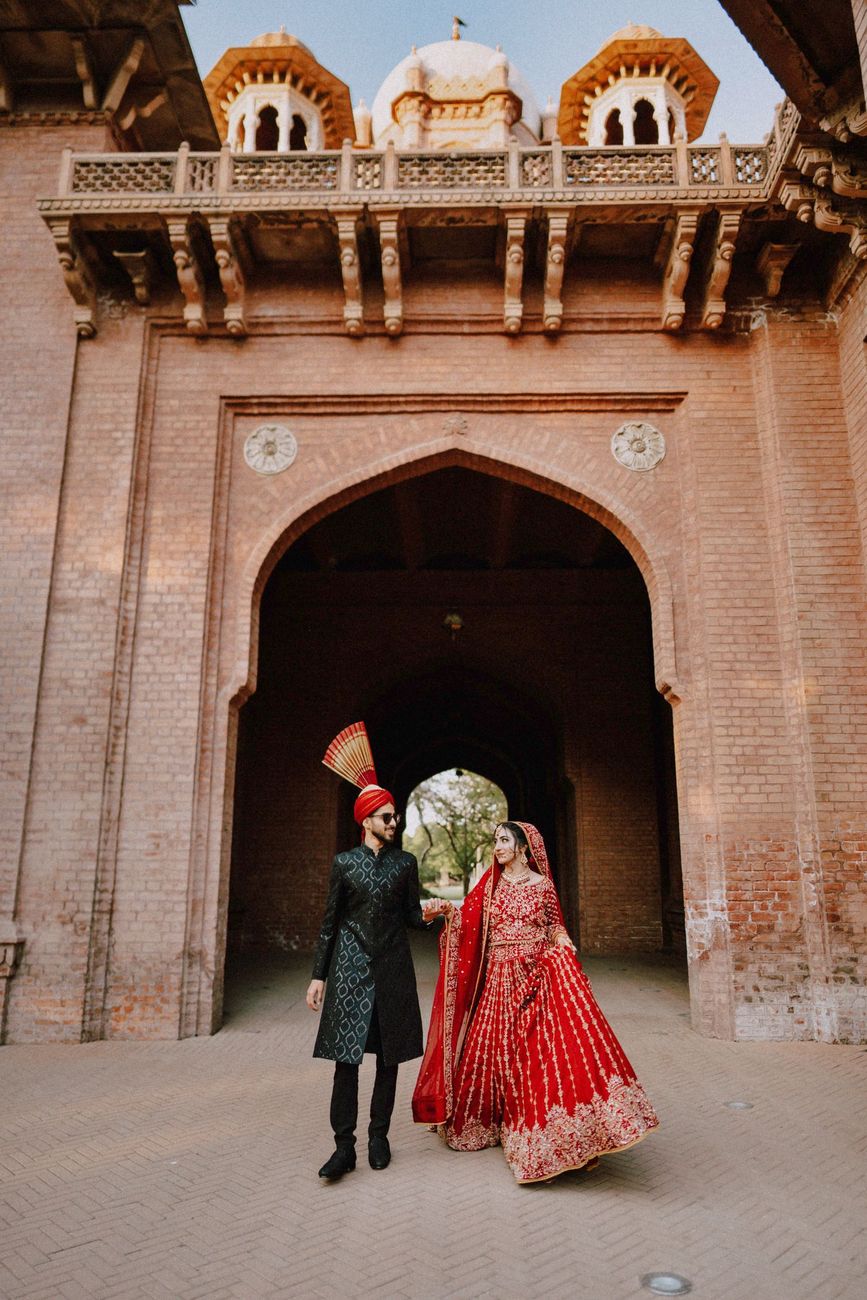 Bride and groom walking hand in hand beneath a grand domed brick gateway, Maham & Aurangzeb's wedding at Aitchison College, Lahore