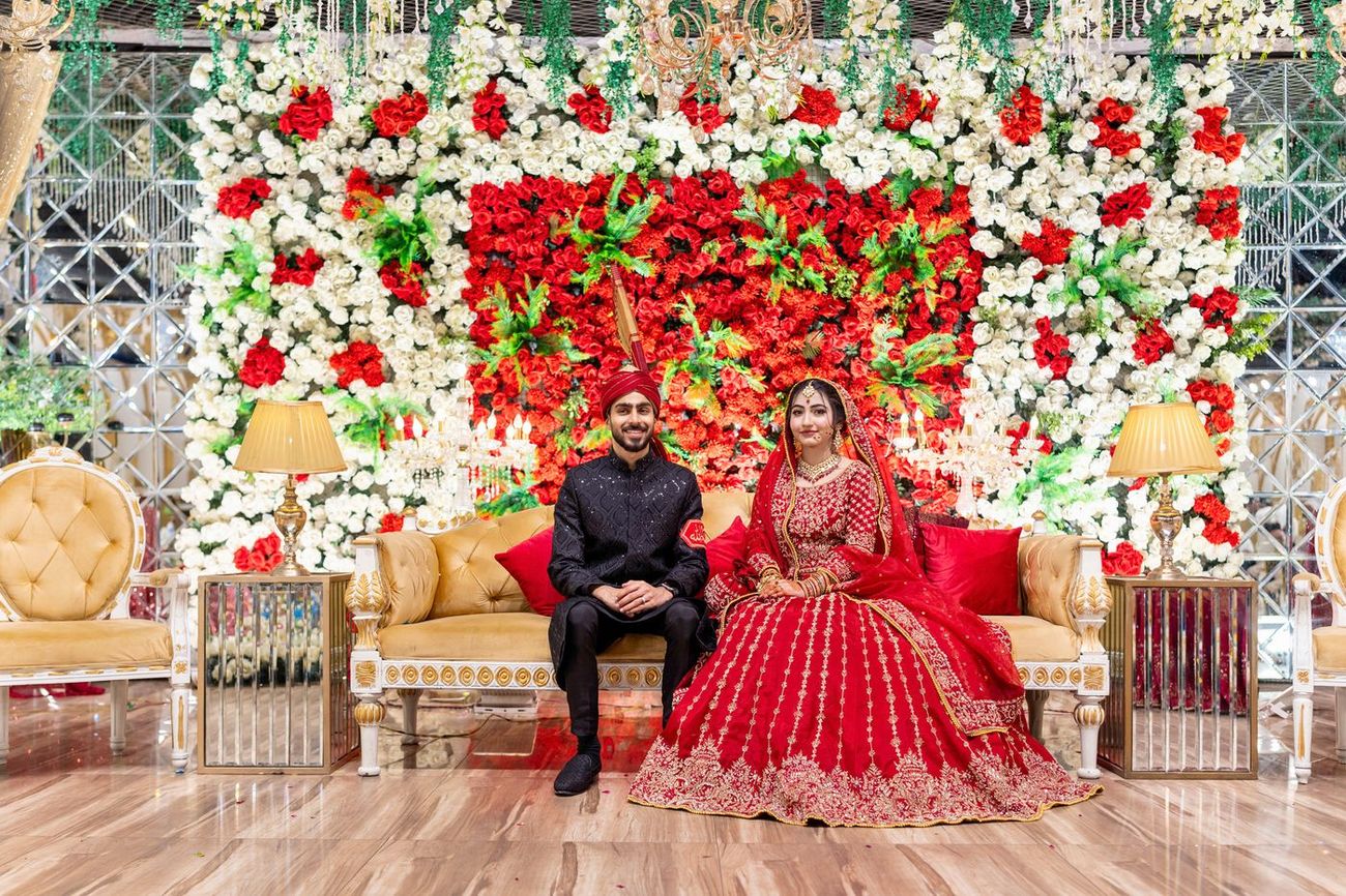 Bride and groom seated together on a gold sofa in front of a red and white flower wall at the reception, Maham & Aurangzeb's wedding