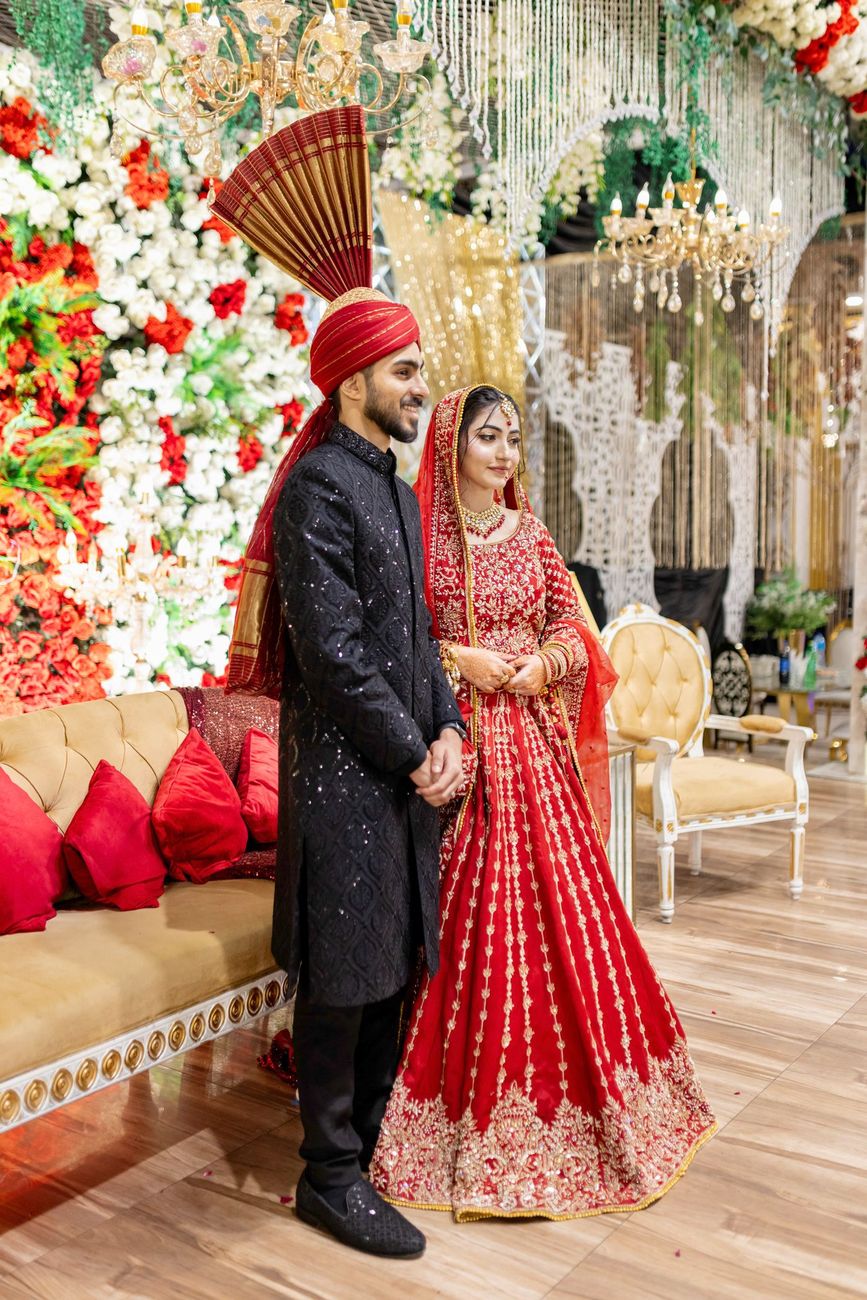 Bride and groom standing together in front of a red and white floral wall and hanging chandeliers, Maham & Aurangzeb's wedding