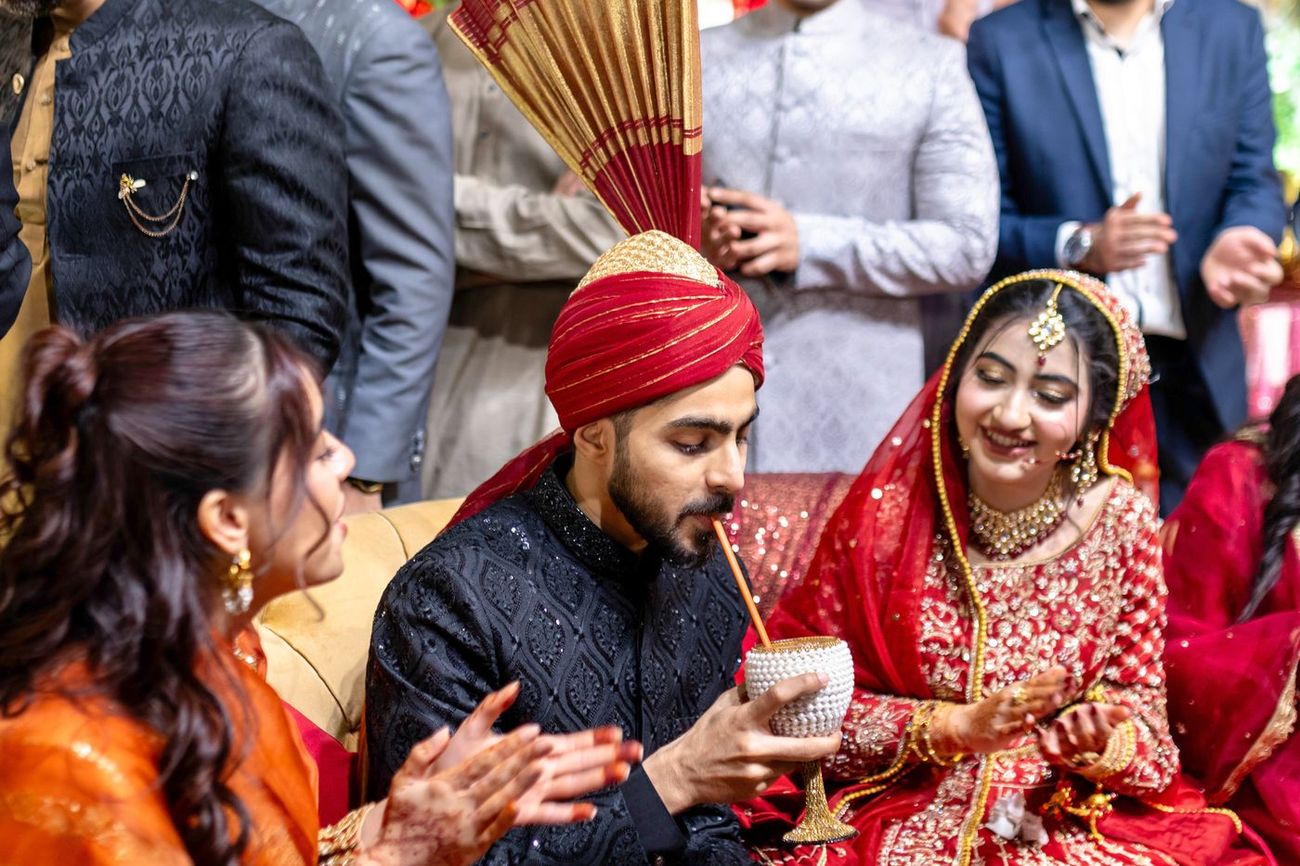 Groom sipping a drink through a straw from a decorated cup as the bride laughs beside him, Maham & Aurangzeb's wedding
