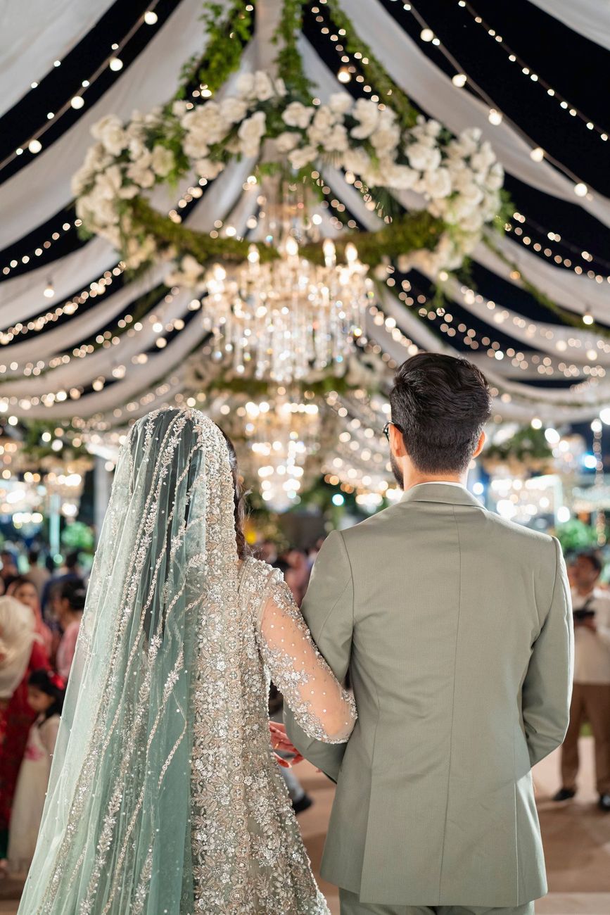 Bride and groom viewed from behind walking beneath floral chandeliers at the walima reception, Maham & Aurangzeb's wedding