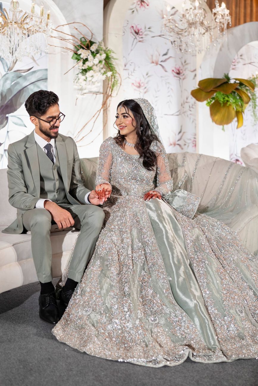 Bride and groom seated together on a sofa at the walima reception, floral arched backdrop behind them, Maham & Aurangzeb's wedding