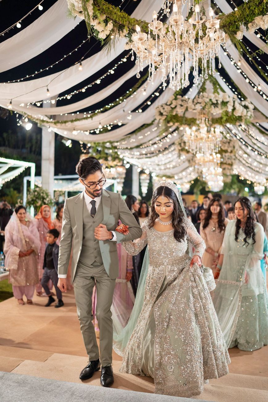 Bride and groom walking together beneath chandeliers and string lights at the walima reception, Maham & Aurangzeb's wedding