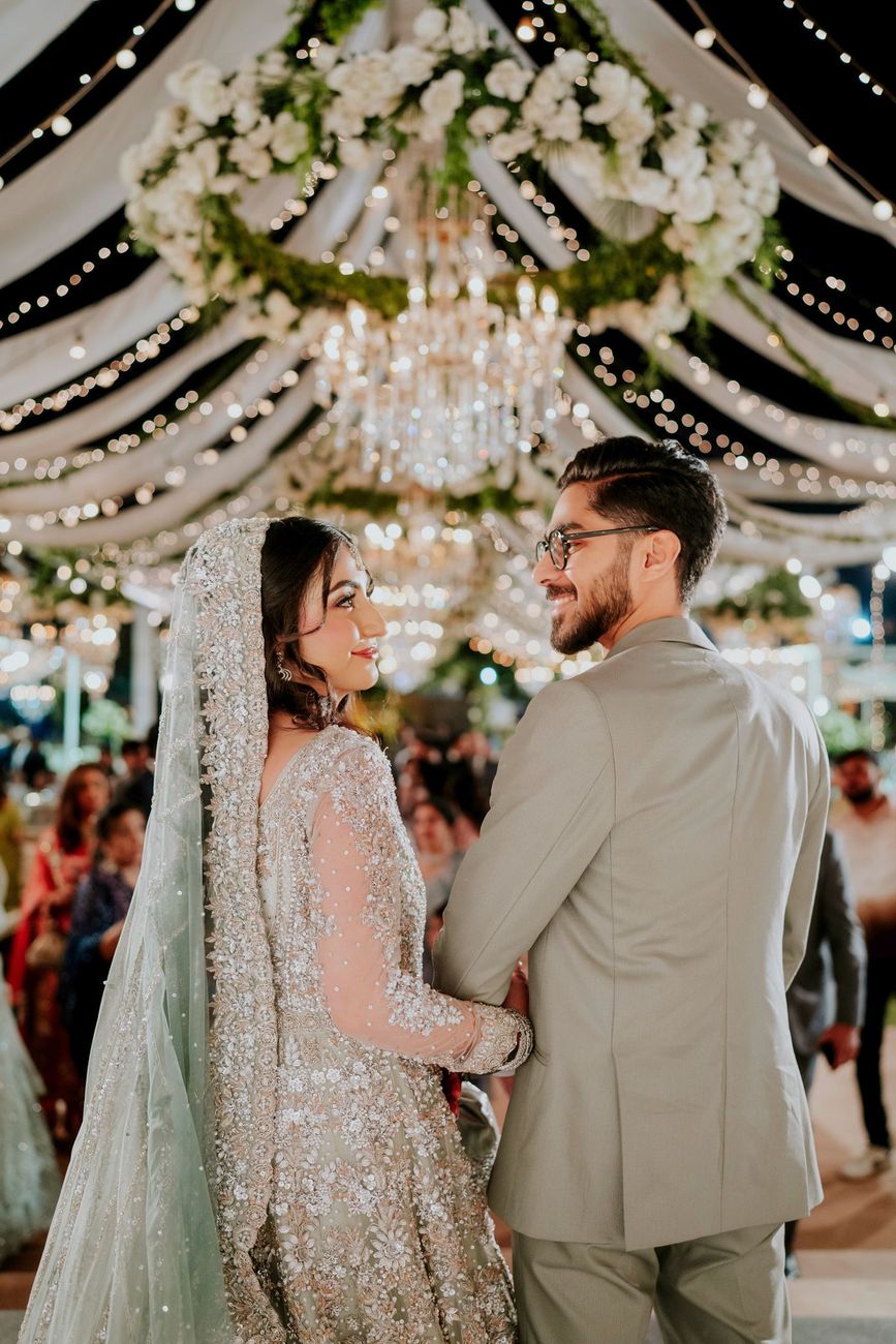 Bride and groom viewed from behind, smiling at each other beneath a floral chandelier at the walima reception, Maham & Aurangzeb's wedding