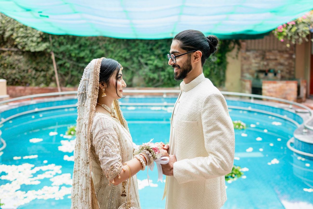 Bride and groom in ivory wedding attire holding hands beside a pool scattered with flower petals, Maham & Aurangzeb's wedding, Lahore