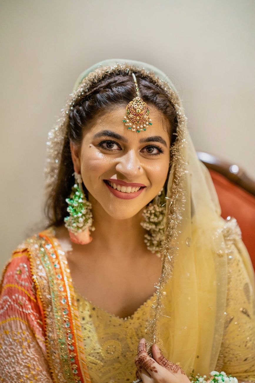 Solo bridal close-up smiling beneath a yellow dupatta, gold jewelry and floral earrings visible, Mashal & Danial's mehndi