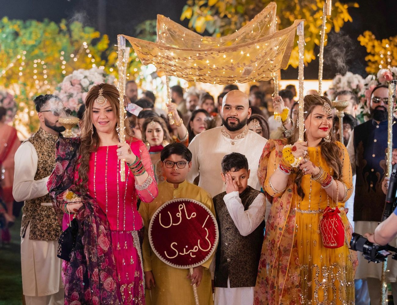 Family members carrying a gold ceremonial canopy with an Urdu sign during the mehndi entrance procession, Mashal & Danial's wedding