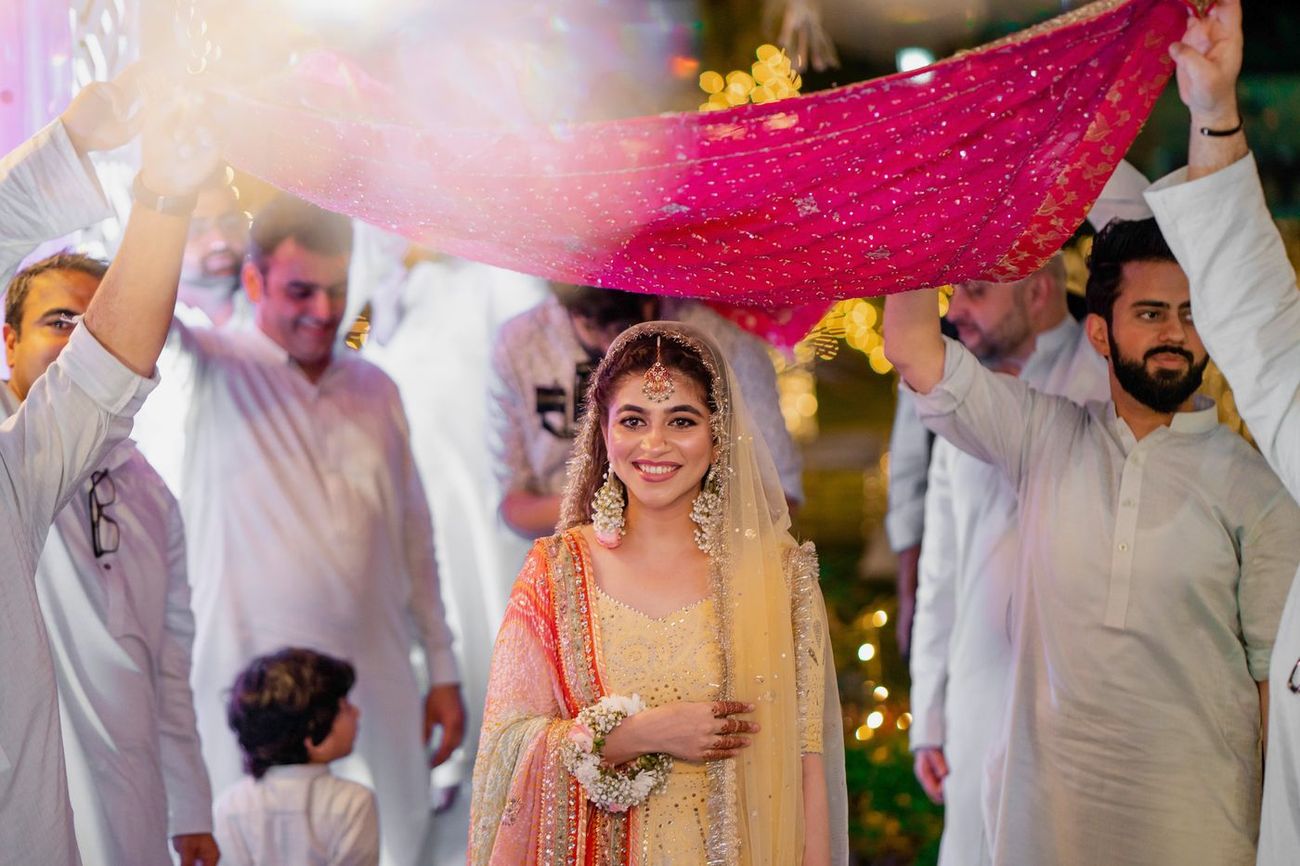 Bride's mehndi entrance beneath a red cloth canopy held up by family, surrounded by photographers, Mashal & Danial's wedding