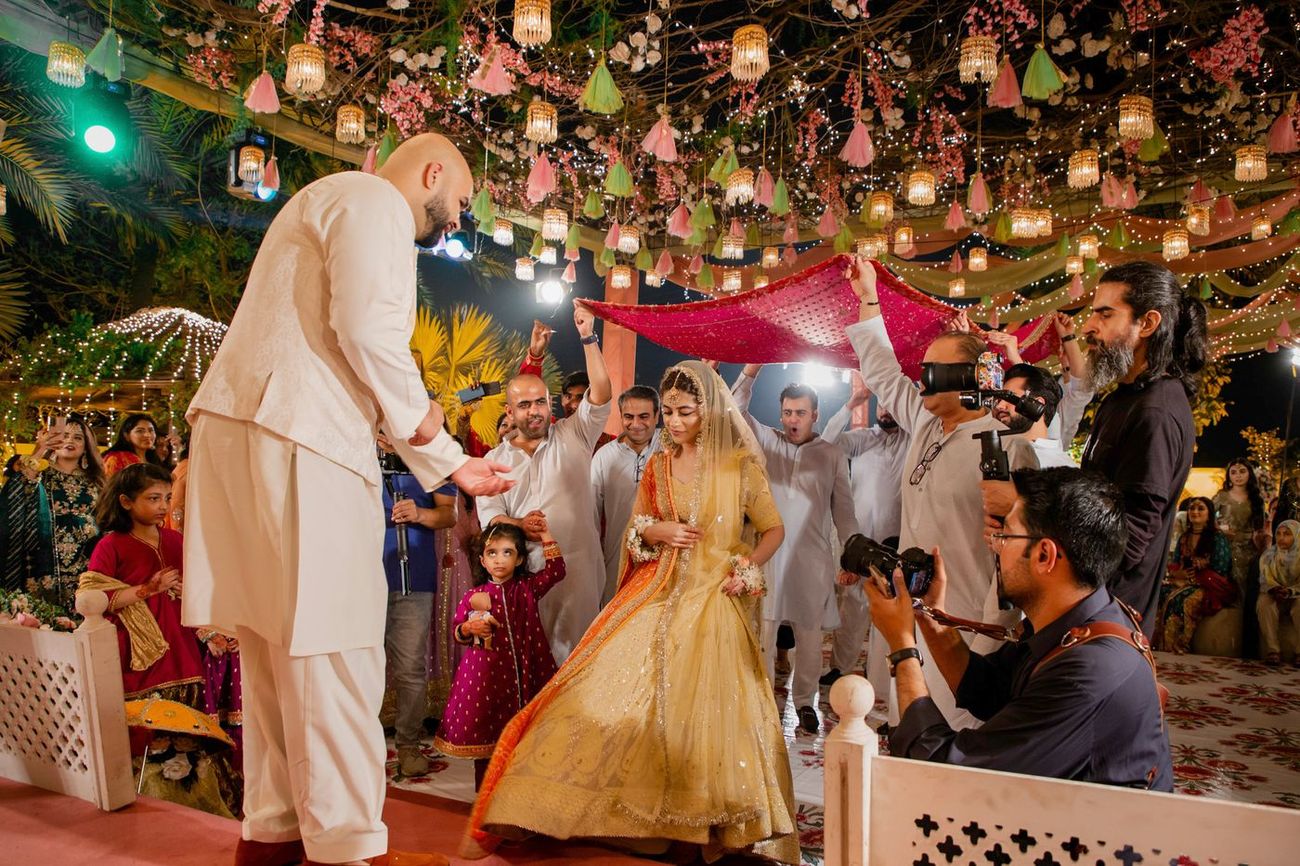 Bride entering the mehndi in a yellow and orange lehenga as the groom waits, family holding a red canopy above her, Mashal & Danial's wedding