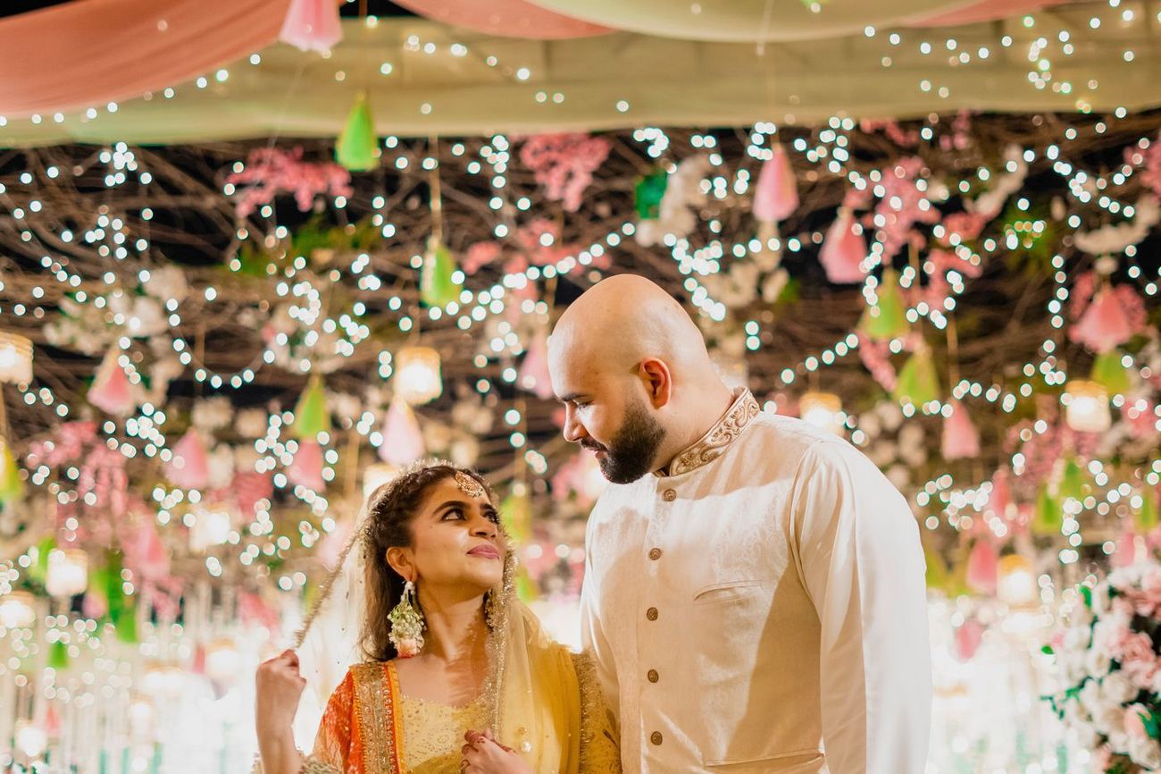 Bride looking up at the groom beneath hanging colorful tassels and lights, Mashal & Danial's mehndi