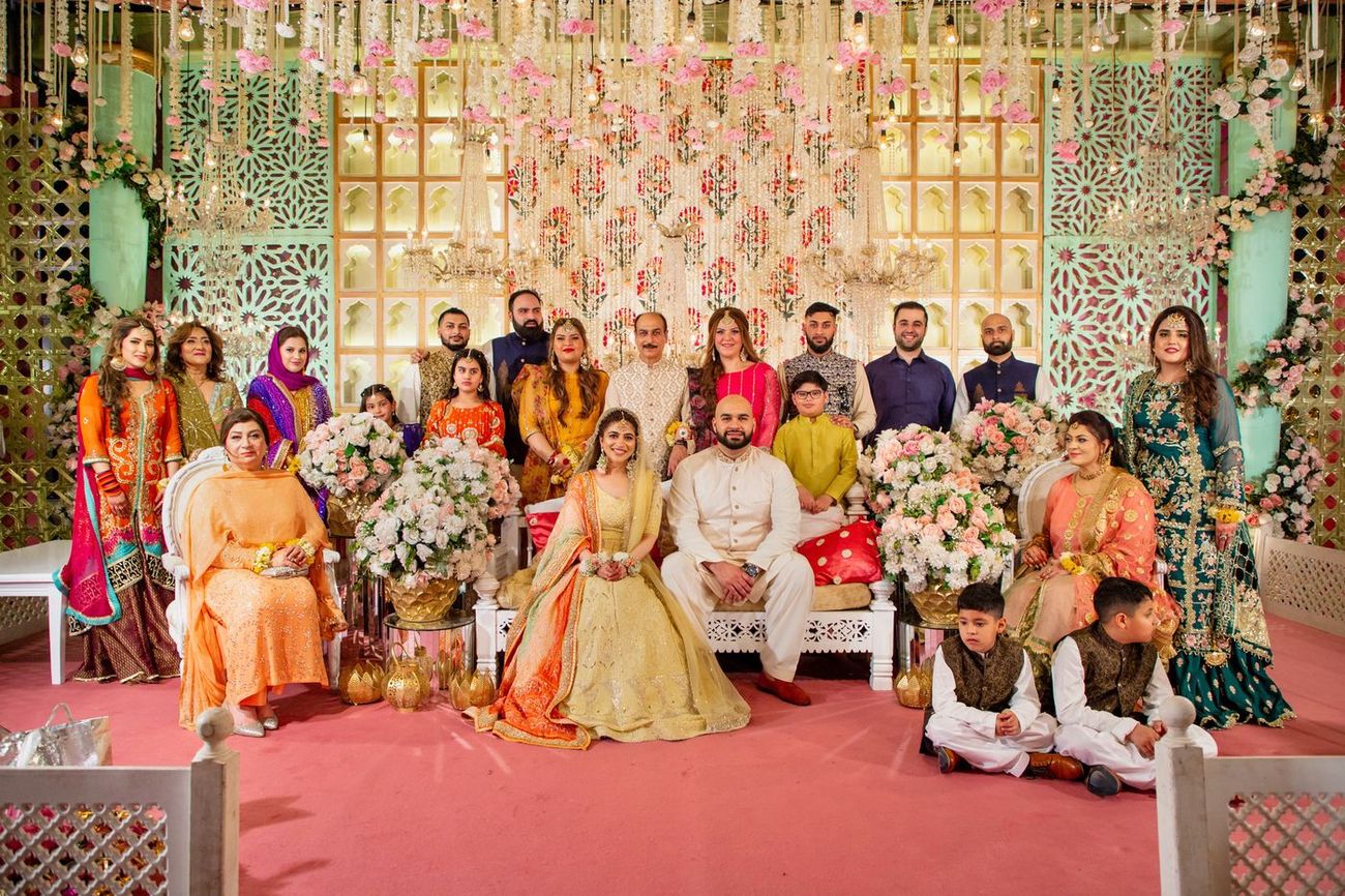 Family group portrait beside the bride and groom on the mehndi stage, floral backdrop behind, Mashal & Danial's wedding