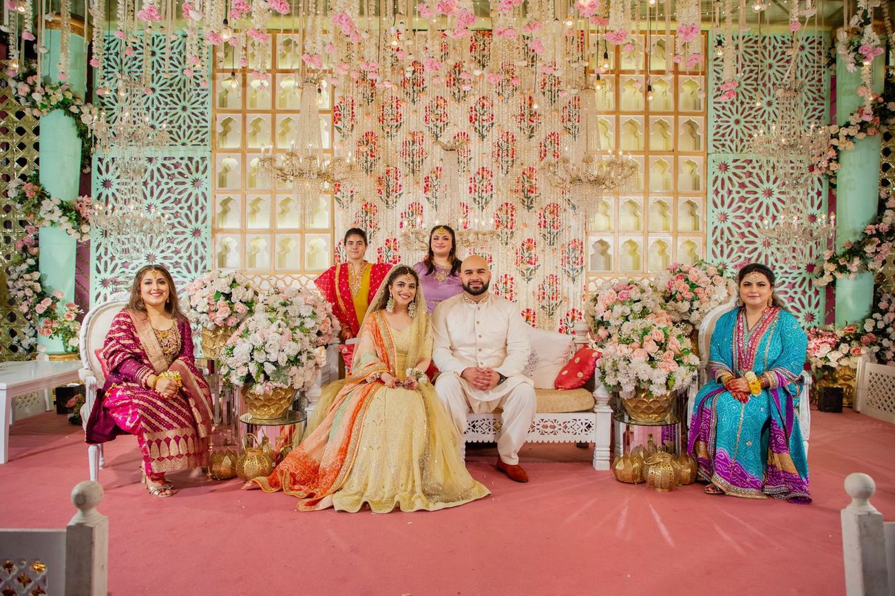 Bride and groom seated with family members on the mehndi stage, gold floral backdrop behind, Mashal & Danial's wedding