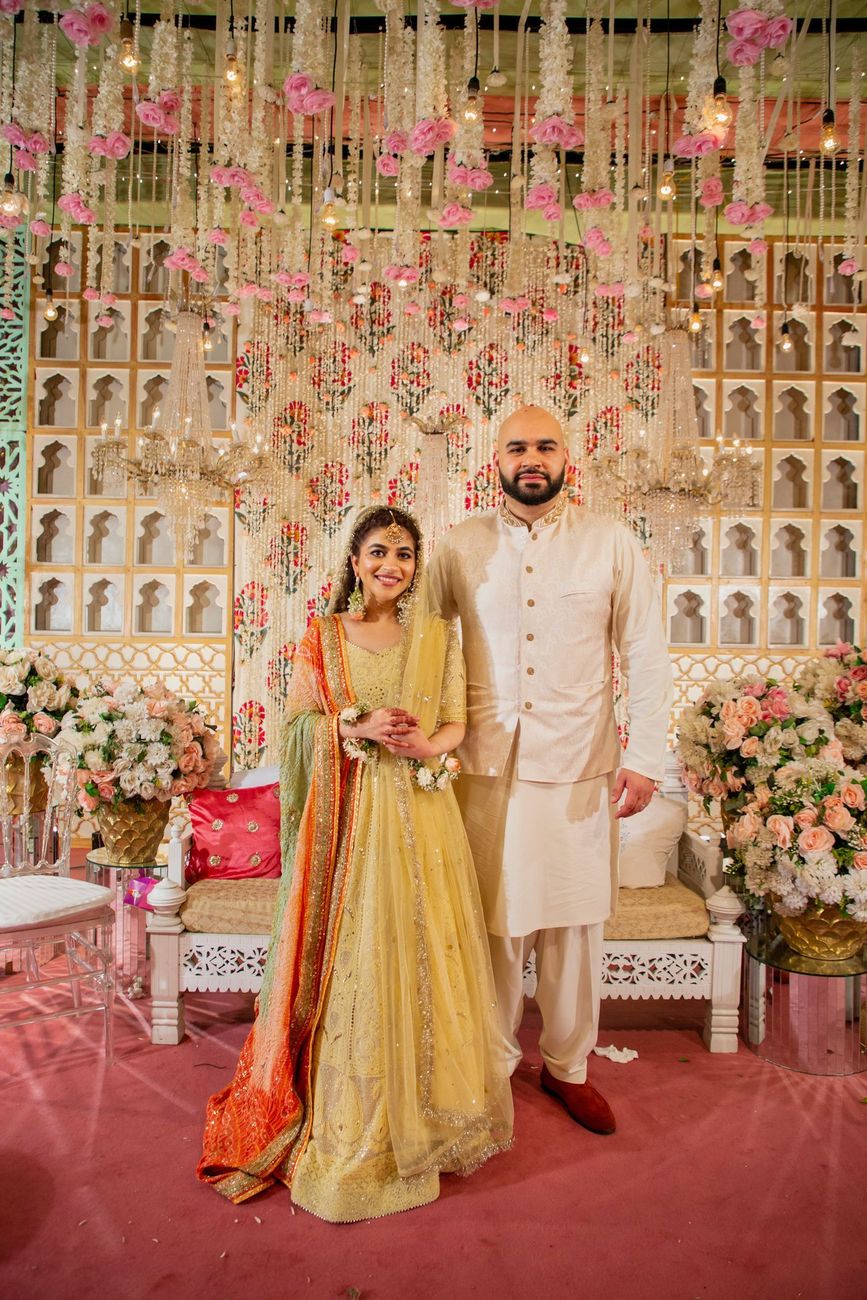 Bride and groom standing together in matching yellow mehndi outfits in front of a floral backdrop, Mashal & Danial's wedding
