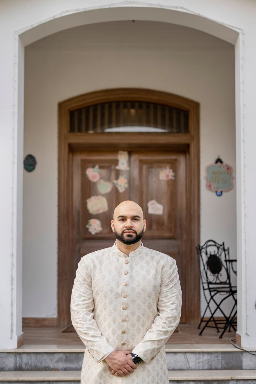 Solo groom portrait standing in a doorway decorated with paper hearts, a Mrs. sign visible beside him, Mashal & Danial's wedding