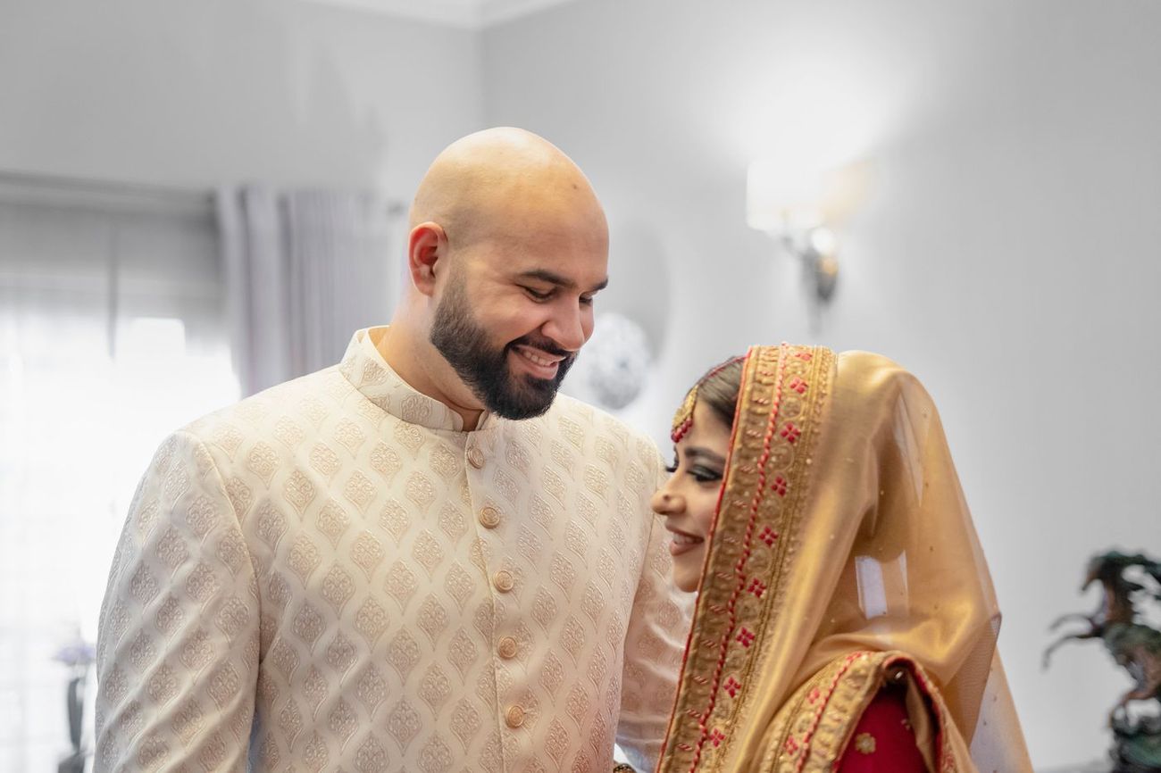Bride and groom smiling together candidly through a doorway, groom in an ivory sherwani and bride in a red and gold outfit, Mashal & Danial's wedding