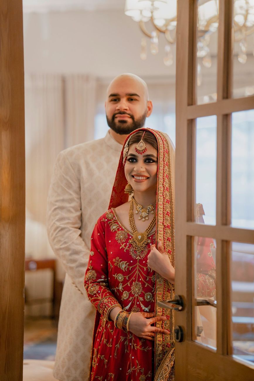 Bride and groom glimpsed through a glass door, bride in a red embroidered outfit smiling at the camera, Mashal & Danial's wedding