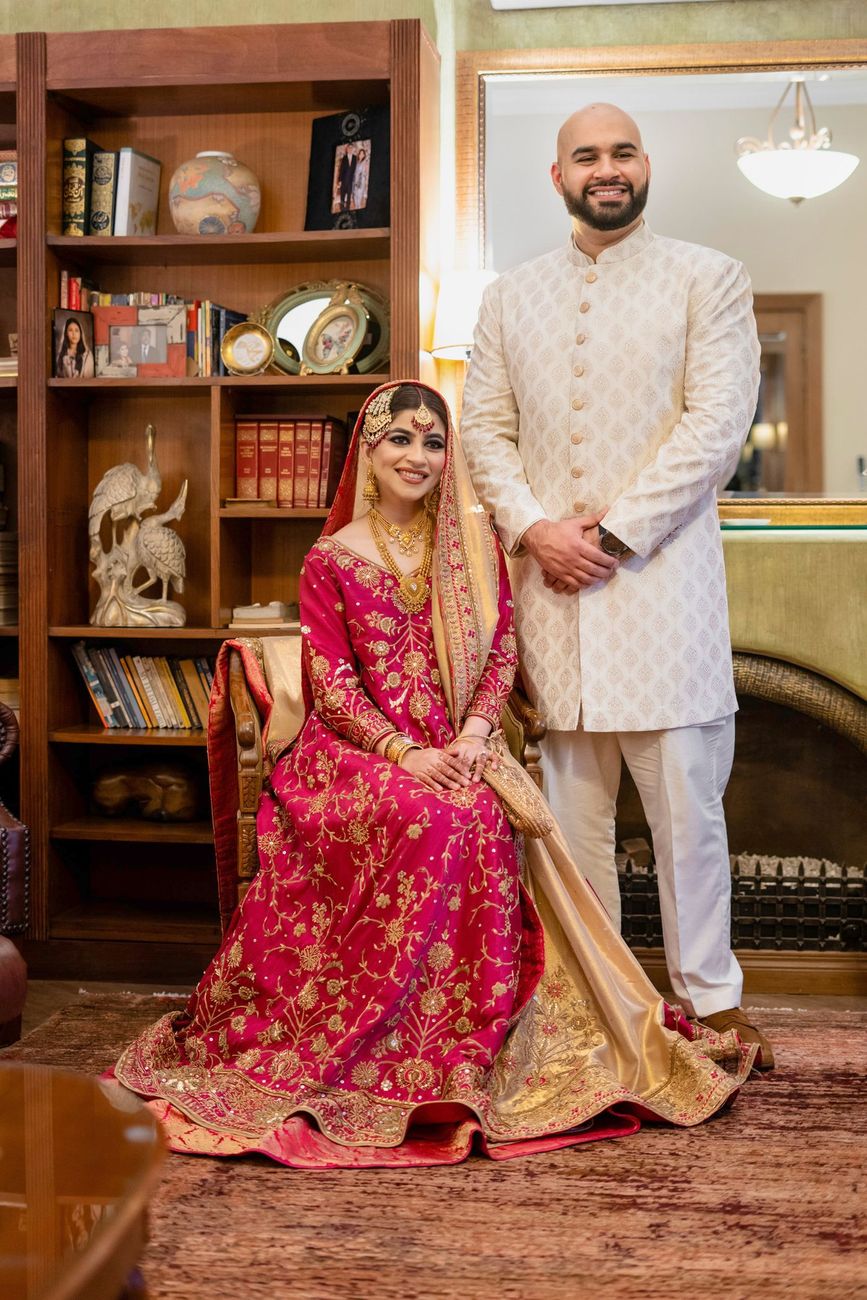Bride seated in a home library with the groom standing beside her, both in red and ivory wedding attire, Mashal & Danial's wedding