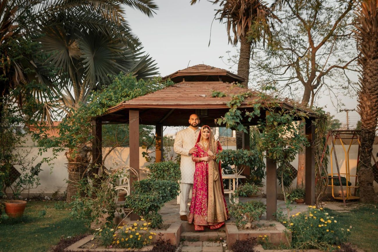 Bride and groom standing together at a garden gazebo, bride in a magenta and gold lehenga, Mashal & Danial's wedding