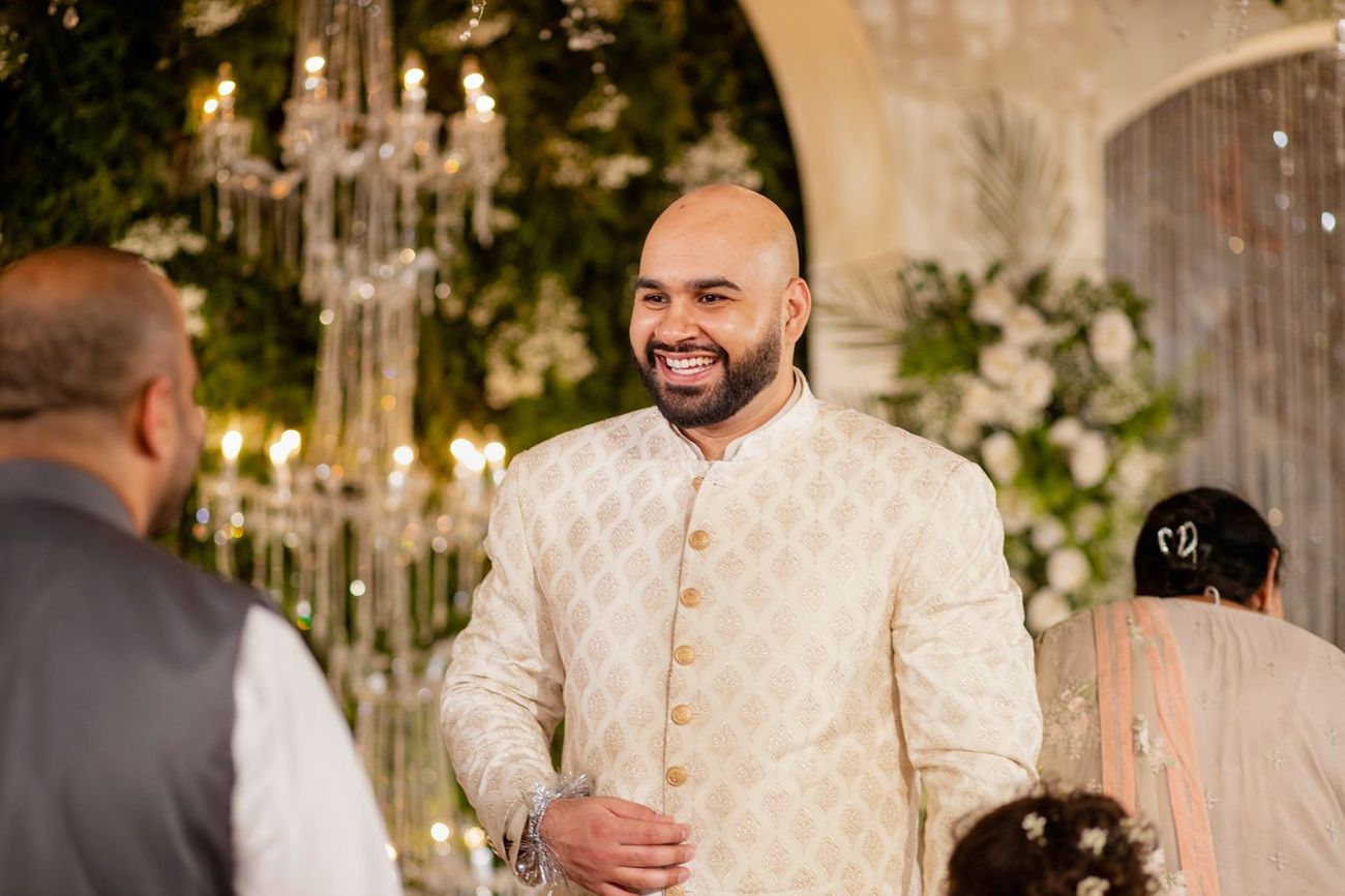 Groom laughing at the reception, crystal chandelier and floral arch visible behind him, Mashal & Danial's wedding