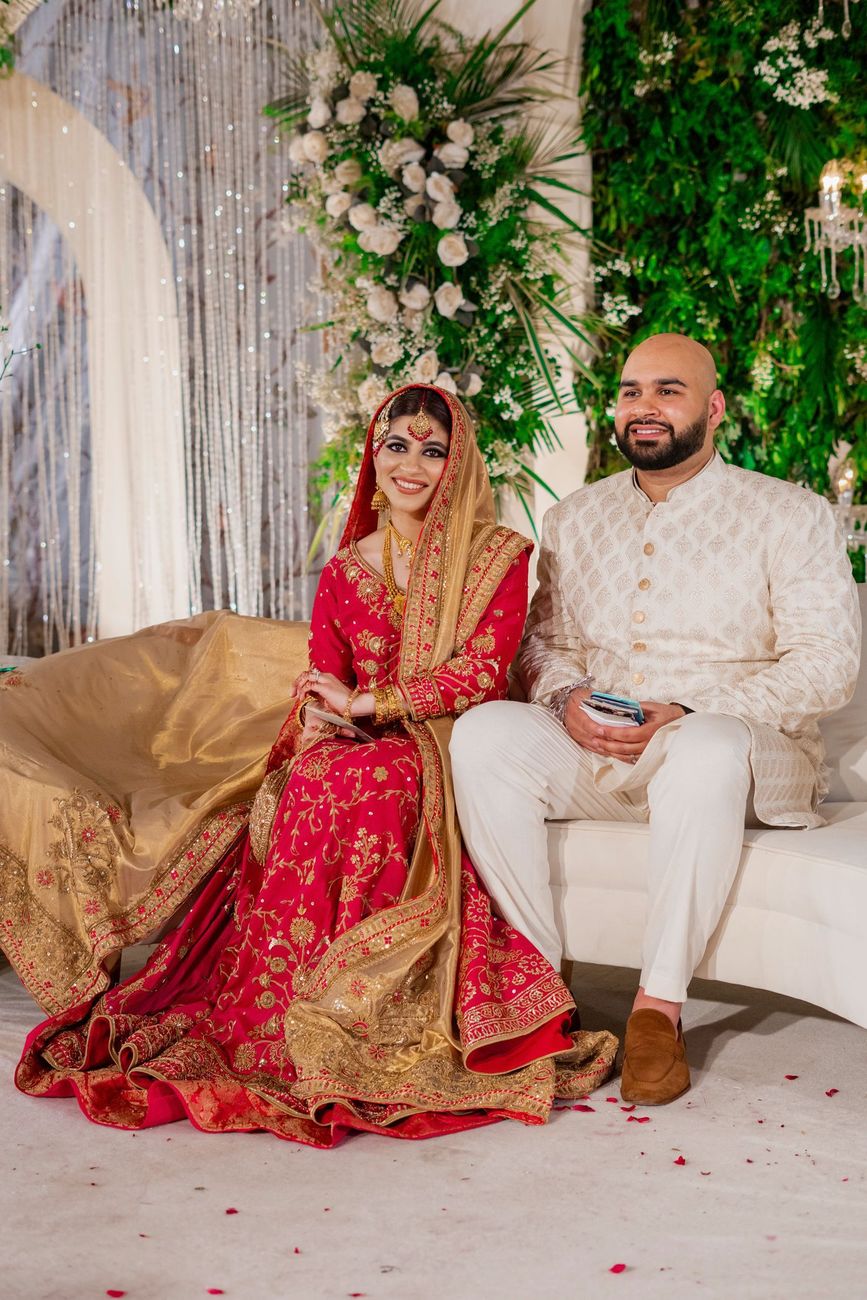 Bride and groom seated together on a white sofa at the reception, bride in a red and gold lehenga, Mashal & Danial's wedding