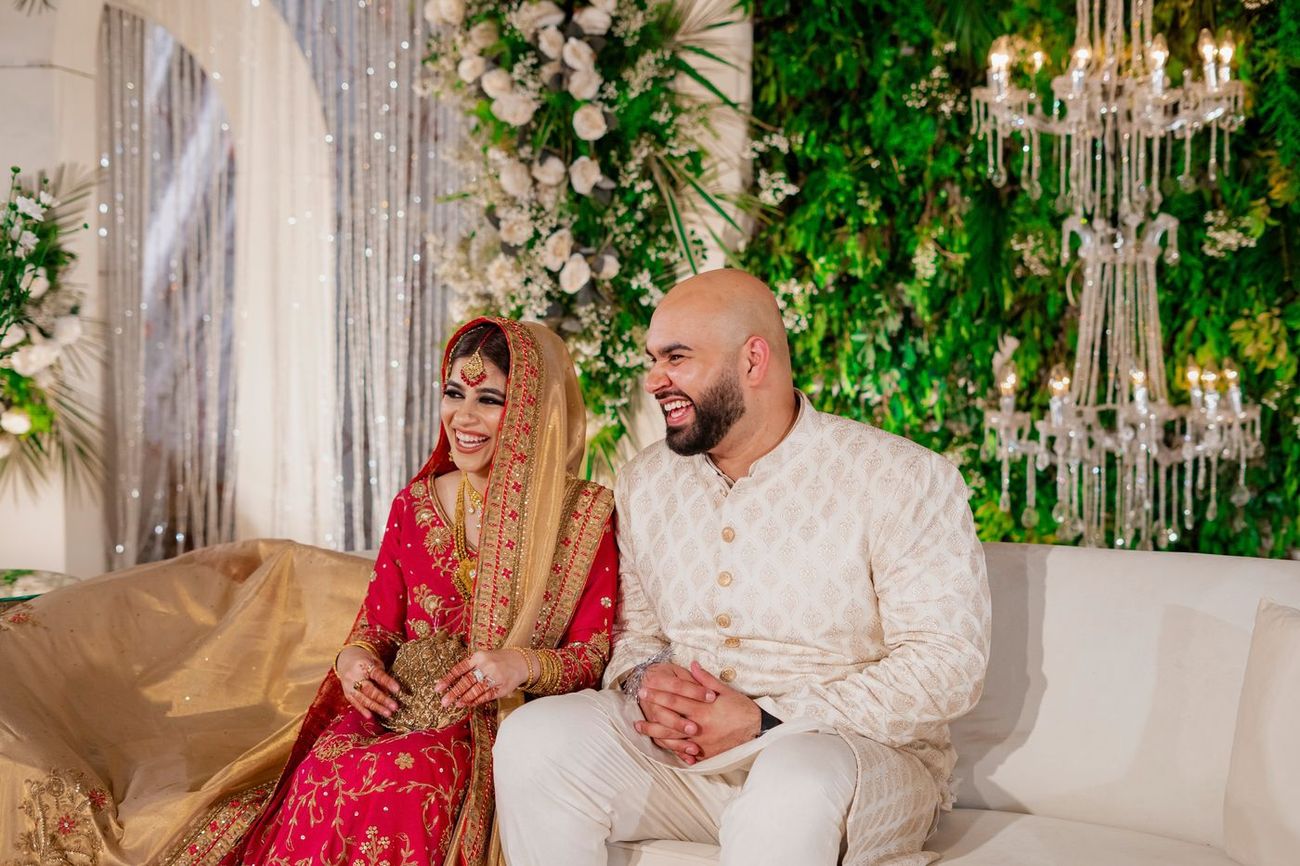 Bride and groom laughing together on a reception sofa, surrounded by greenery and chandeliers, Mashal & Danial's wedding