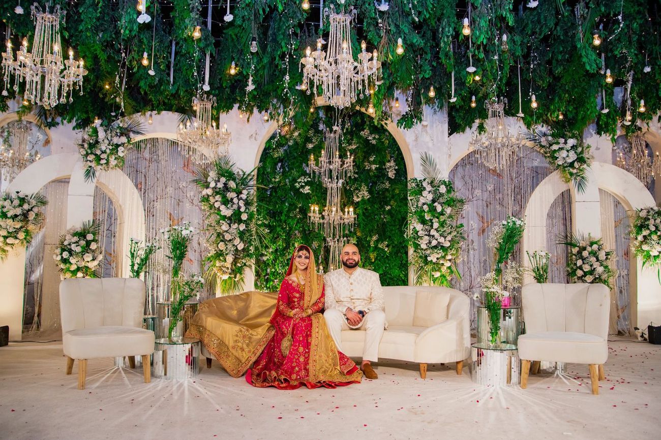 Bride and groom seated together on the reception stage framed by arches and hanging greenery, Mashal & Danial's wedding